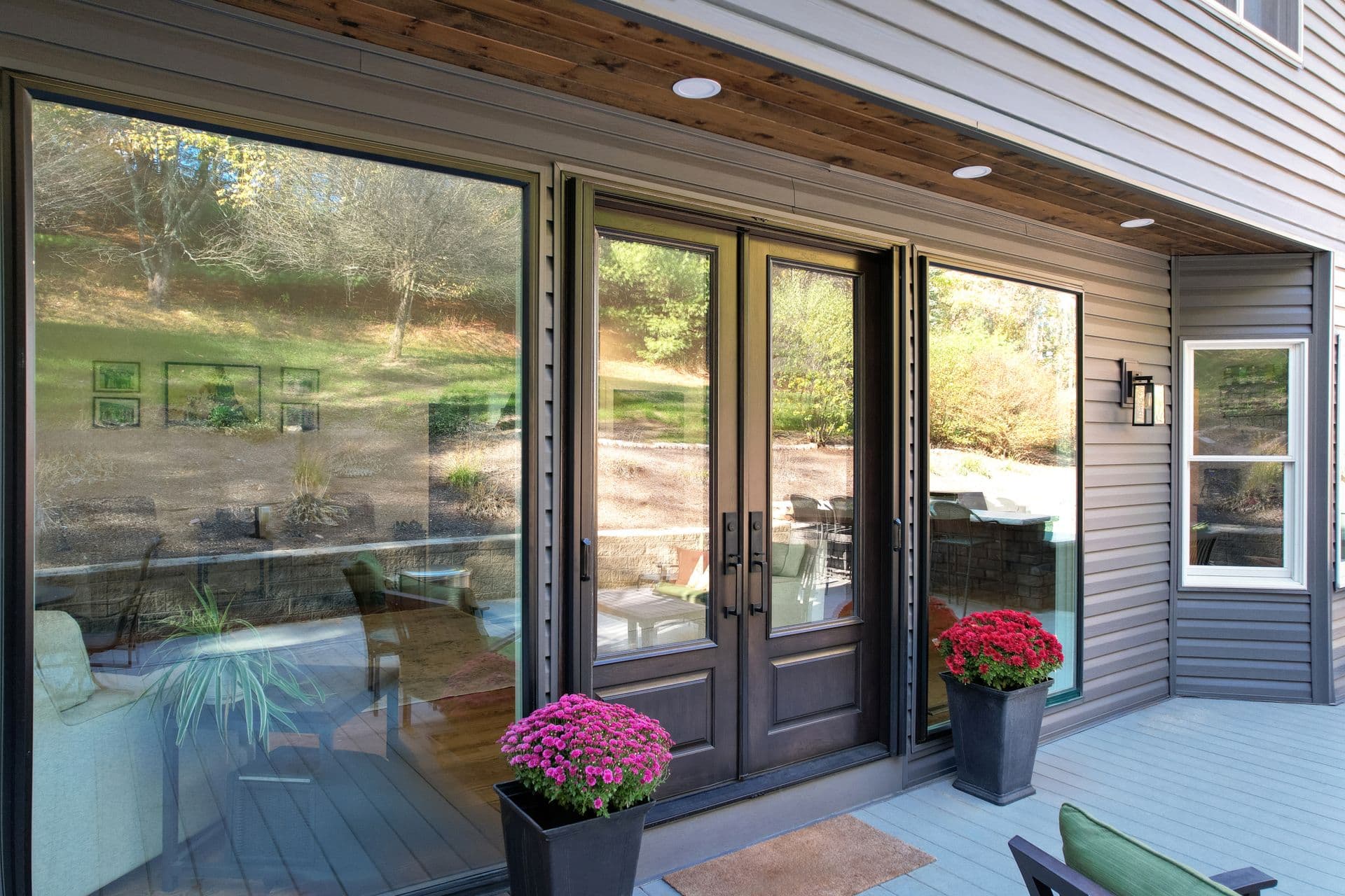 Covered patio with large glass doors and windows, pink mums in planters, wooden deck, and forested landscape view