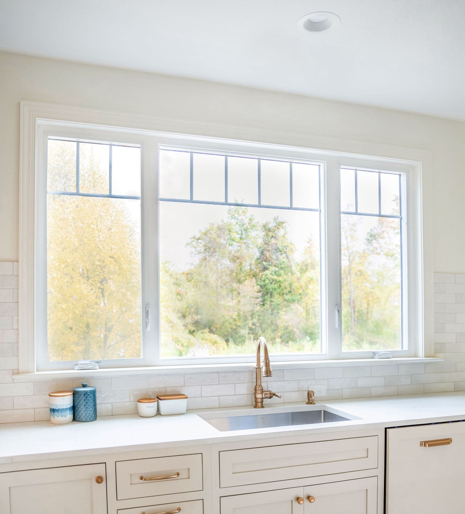 Kitchen sink with large windows overlooking trees and autumn foliage, brass faucet, white countertop, and cream-colored cabinetry with brass hardware