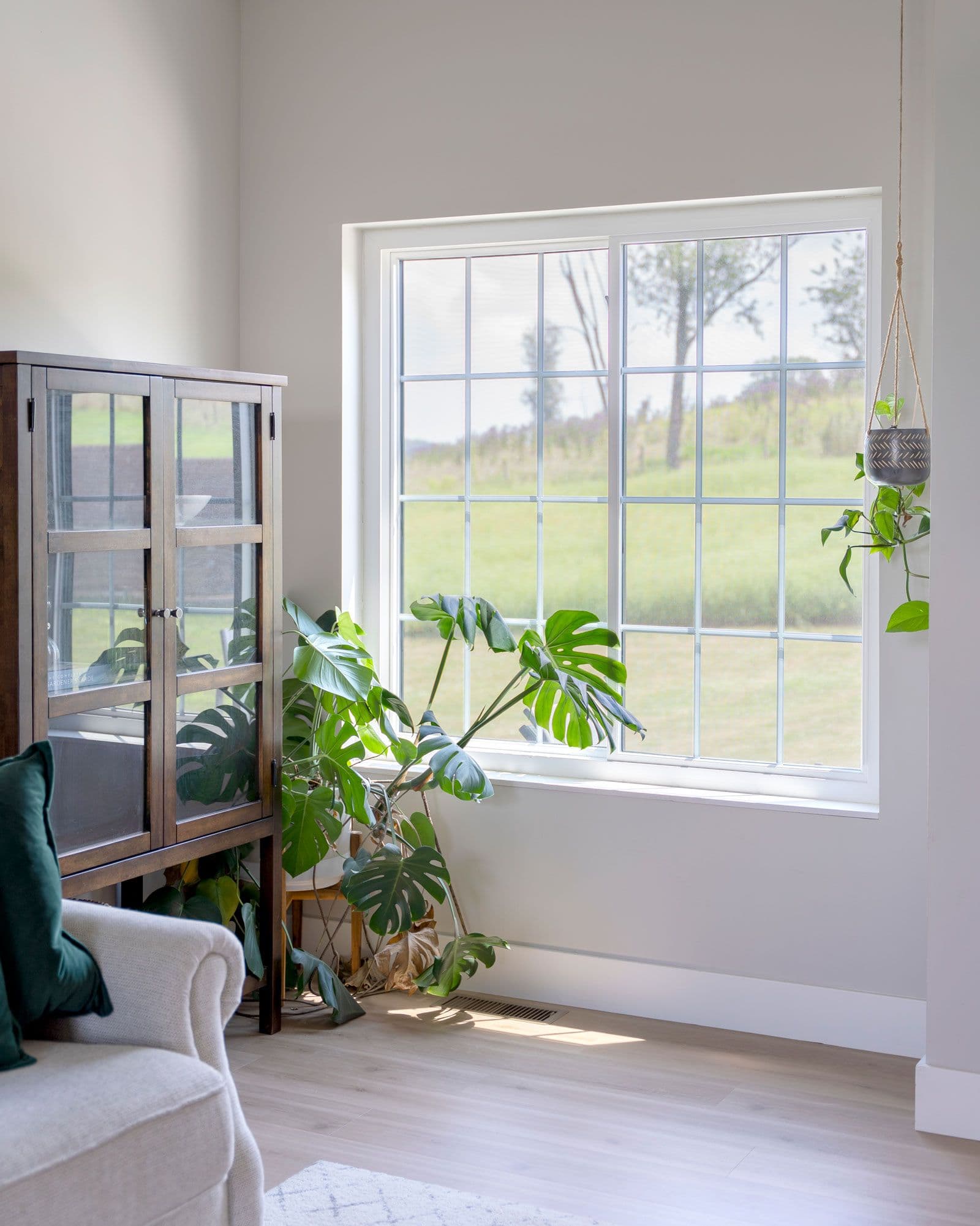 Bright living room with large multi-pane windows overlooking green fields, potted plants on windowsill and hanging basket, upholstered chair with dark green pillow, and glass display cabinet with interior plants