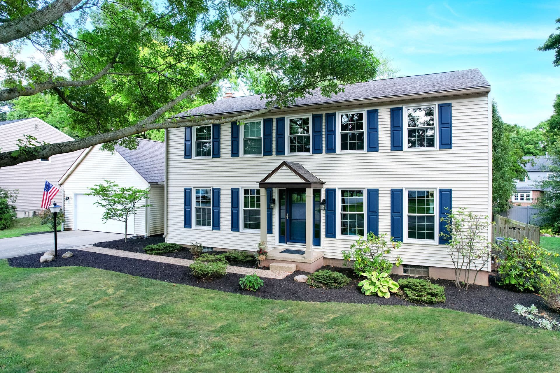 White two-story colonial home with blue shutters and trim, front porch entry, landscaped yard with large tree overhead, and attached garage with American flag