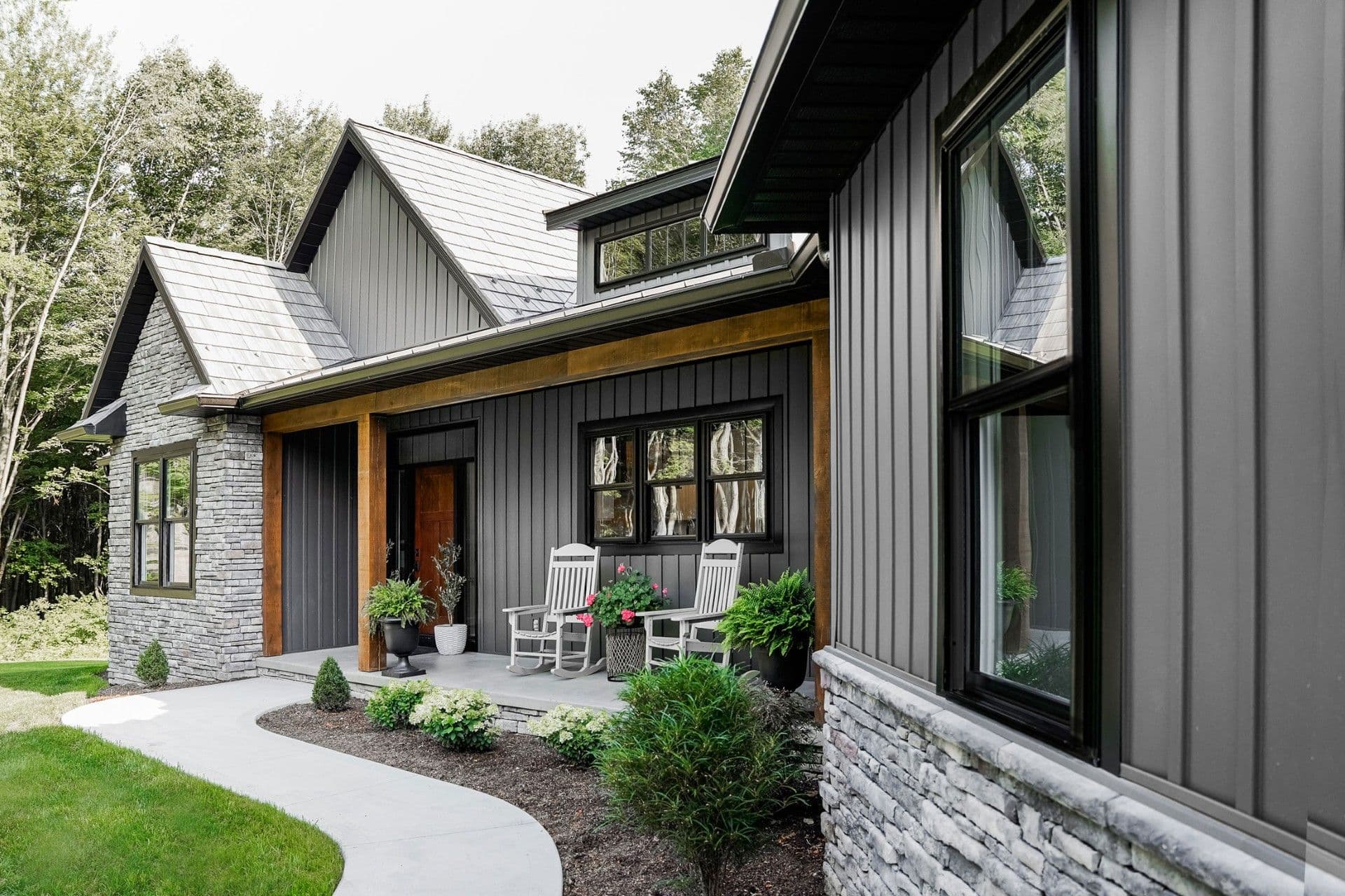 Modern farmhouse exterior with black metal siding, white stone accents, metal roofing, and wooden entry overhang, featuring a patio with white Adirondack chairs surrounded by landscaping and trees