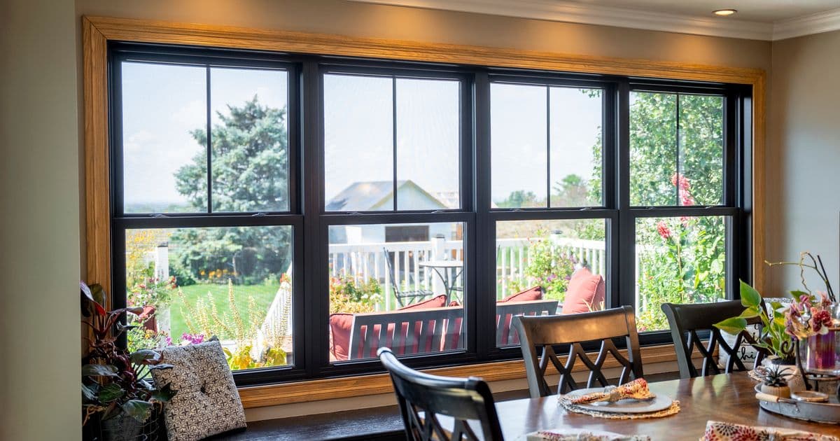 Large black-framed sliding glass doors with wood trim open onto a patio and garden view, with dining chairs and potted plants on the windowsill