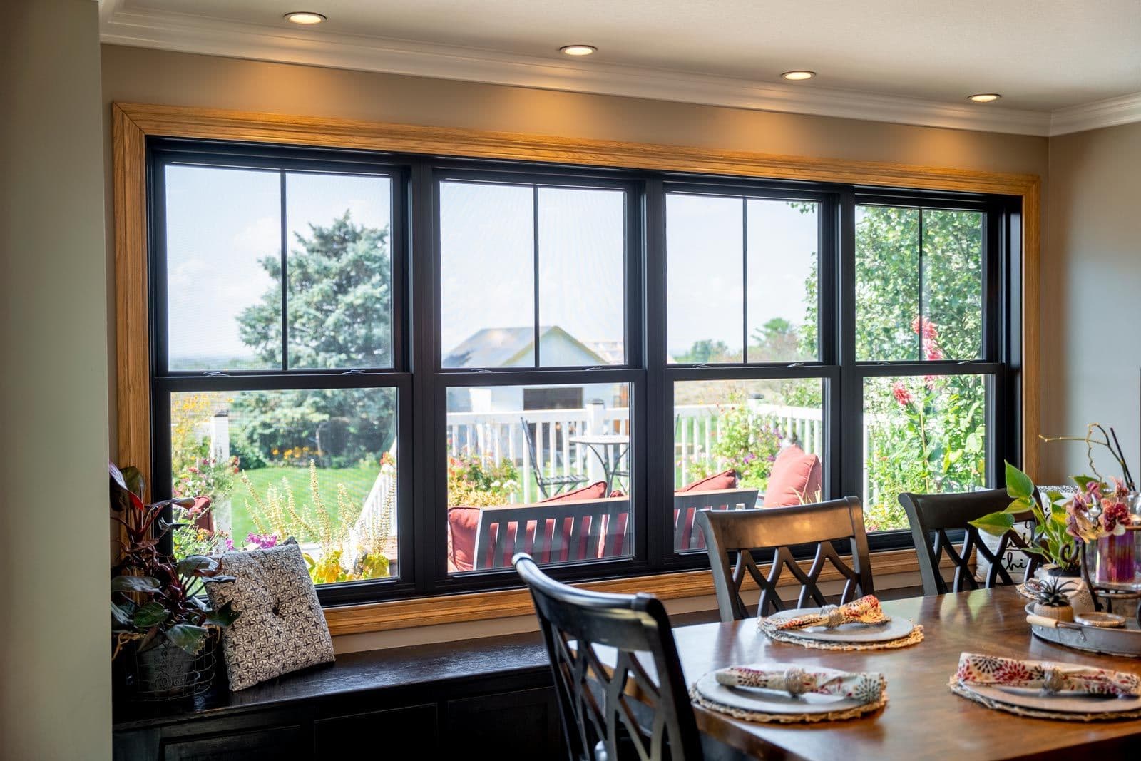 Dining room with black-framed sliding glass doors opening to backyard patio with garden and lounge seating, wooden window seat with cushion, table set for dining in foreground
