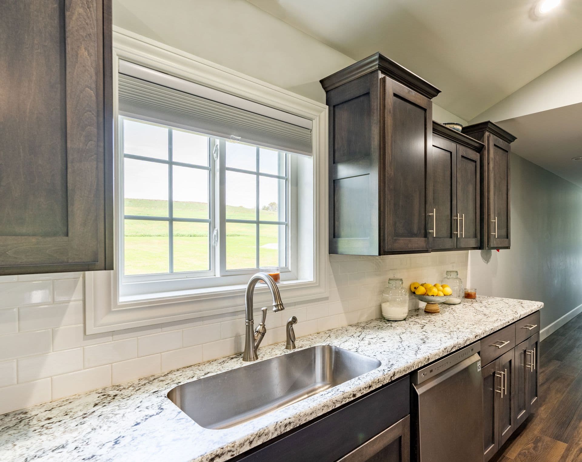 Modern kitchen with stainless steel sink, dark wood cabinets with glass doors, granite countertop, white subway tile backsplash, and large window overlooking green landscape