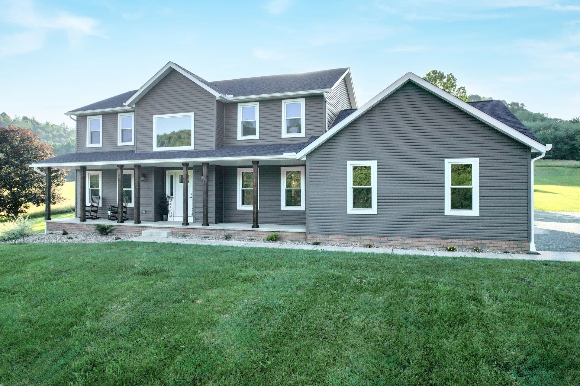 Modern two-story farmhouse with gray siding, white trim, front porch with columns, brick foundation, and manicured lawn surrounded by green fields