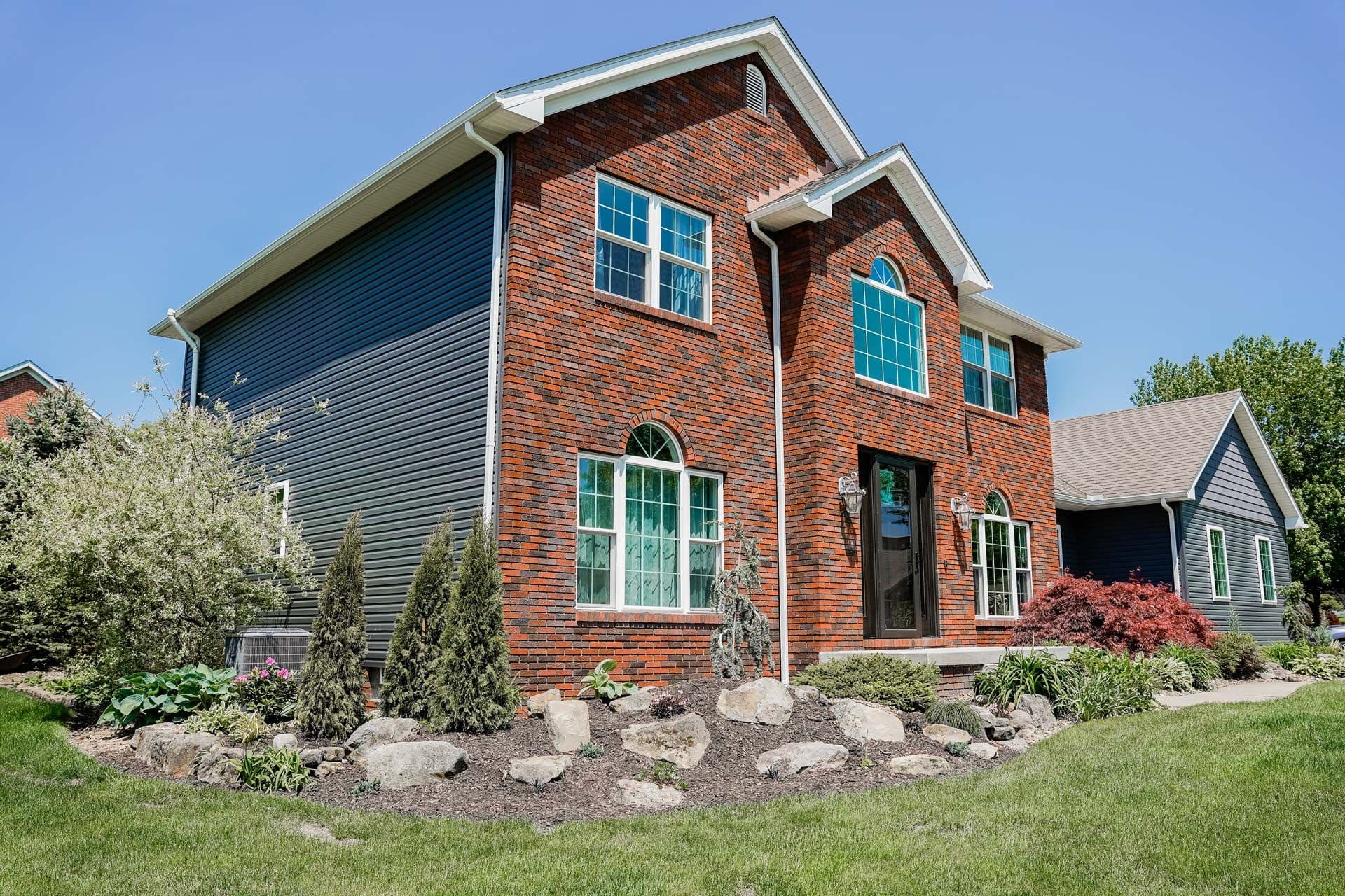 Two-story brick home with attached garage, blue-trimmed windows, landscaped front yard with stone accents and shrubs under clear sky