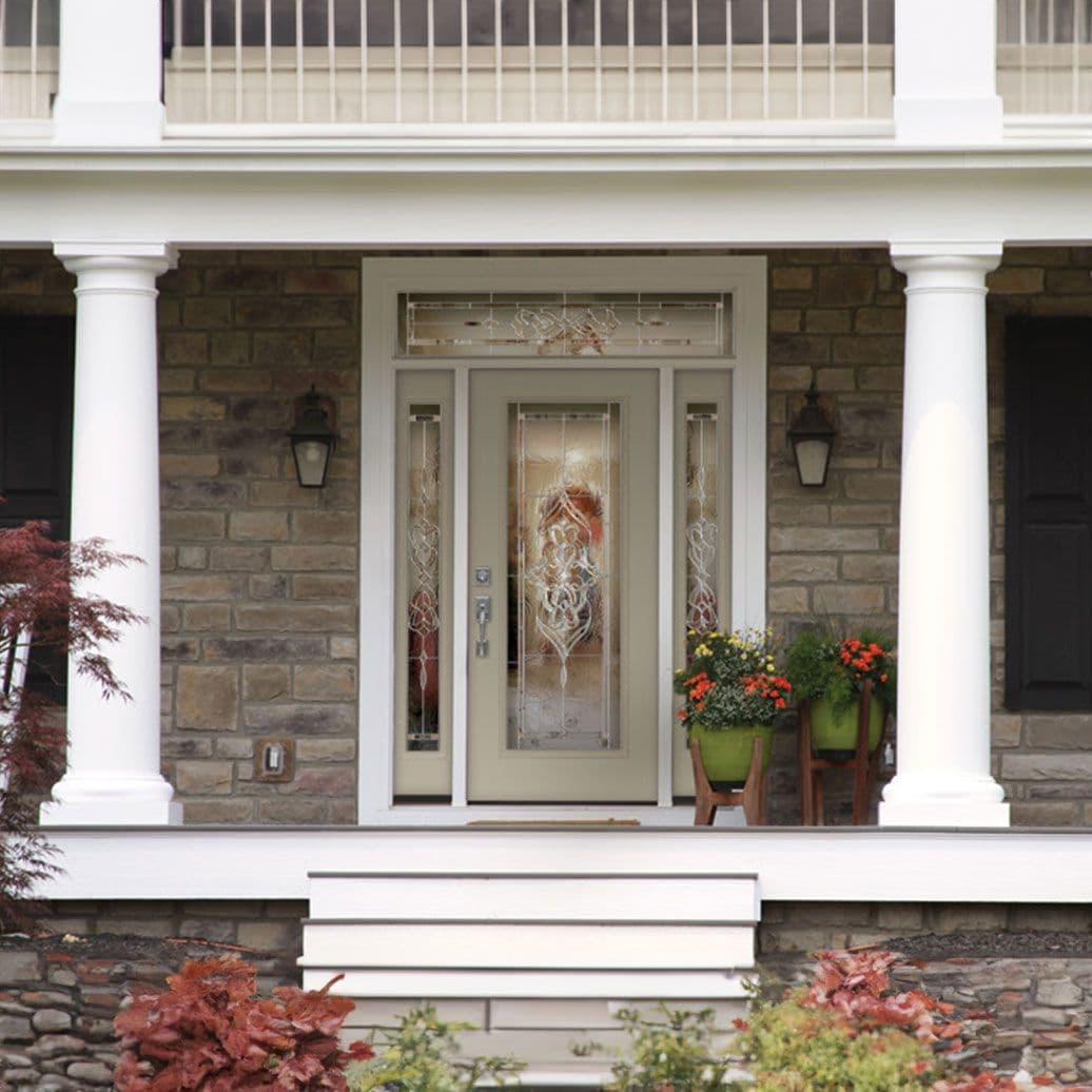 Classic front entrance with cream-colored door featuring glass panels with decorative wreath, flanked by white columns, stone facade, lantern sconces, potted flowers, and white steps