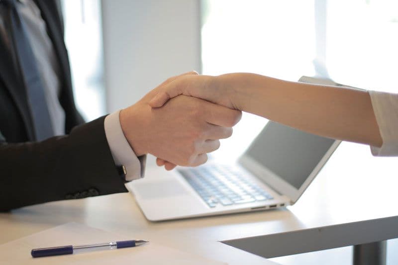 Two business professionals shaking hands over a desk with laptop and paperwork, sealing a deal or agreement