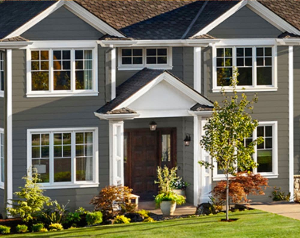 Modern two-story gray house with white trim, dark roof, multiple windows, dark red front door, landscaped entry with plants and small trees, and manicured lawn