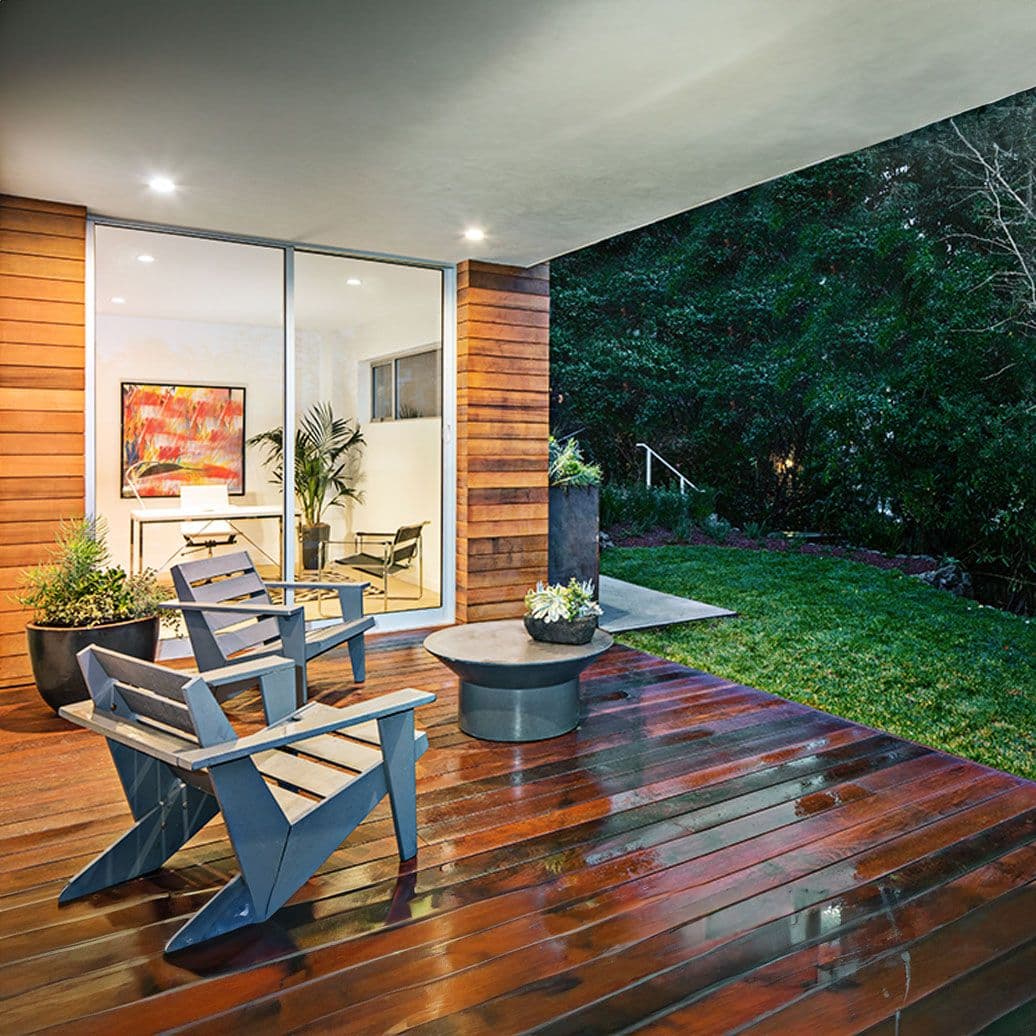 Modern patio with blue Adirondack chairs on wet wooden deck, potted plants, round fire table, and interior view of contemporary home with artwork and green wall backdrop at night