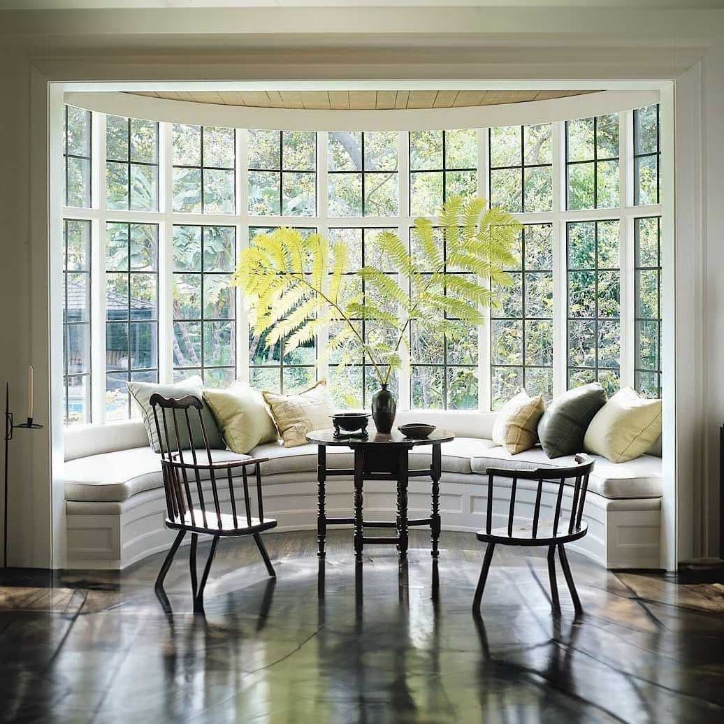 Bay window seating area with curved white cushioned bench, two black chairs facing a small black table, yellow fern plant, and large multi-pane windows overlooking greenery