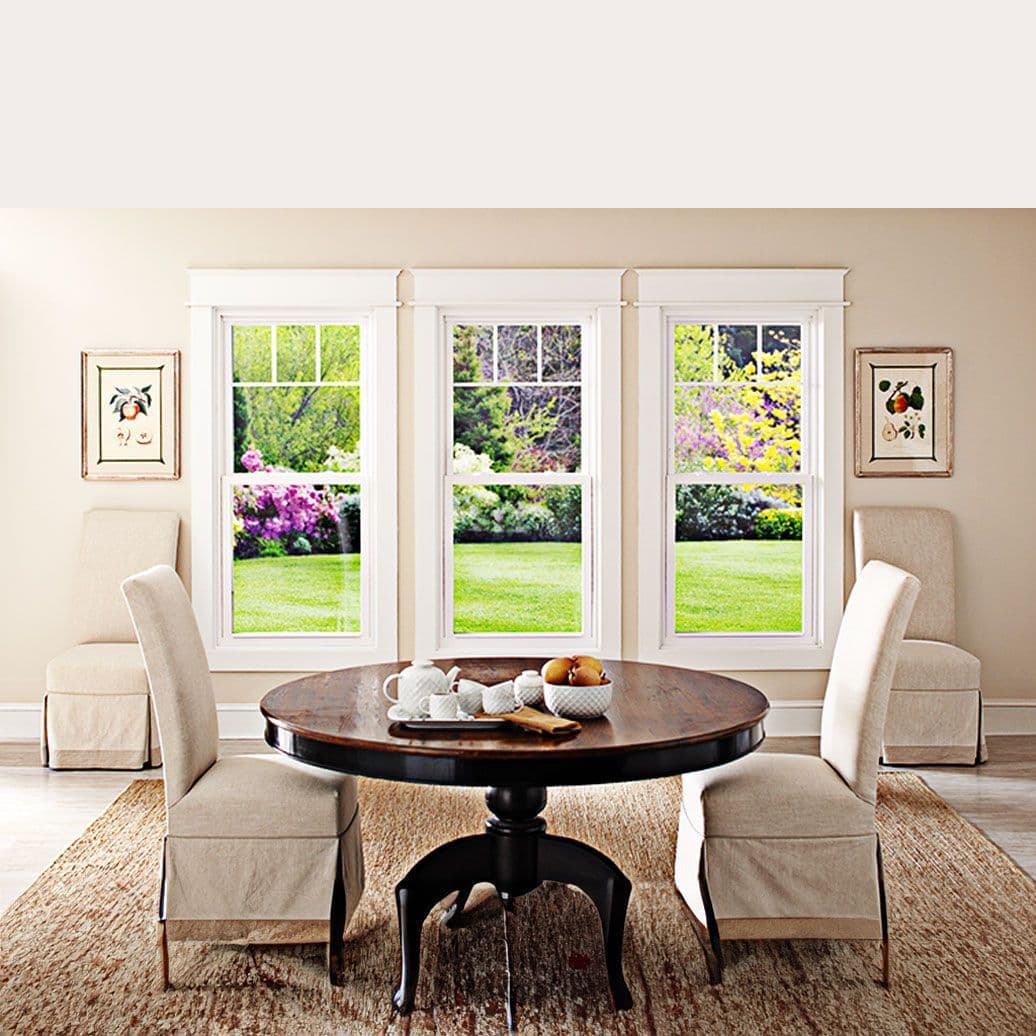 Bright living room with cream-colored chairs around a dark round pedestal table, three large windows showing blooming garden, and botanical artwork on beige walls