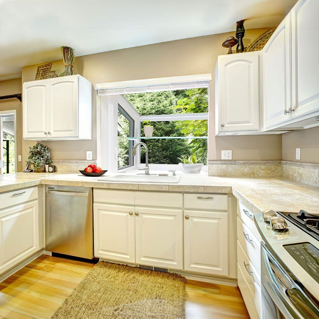Bright kitchen with white cabinetry, beige countertops, stainless steel appliances, and a large window overlooking green foliage above the sink