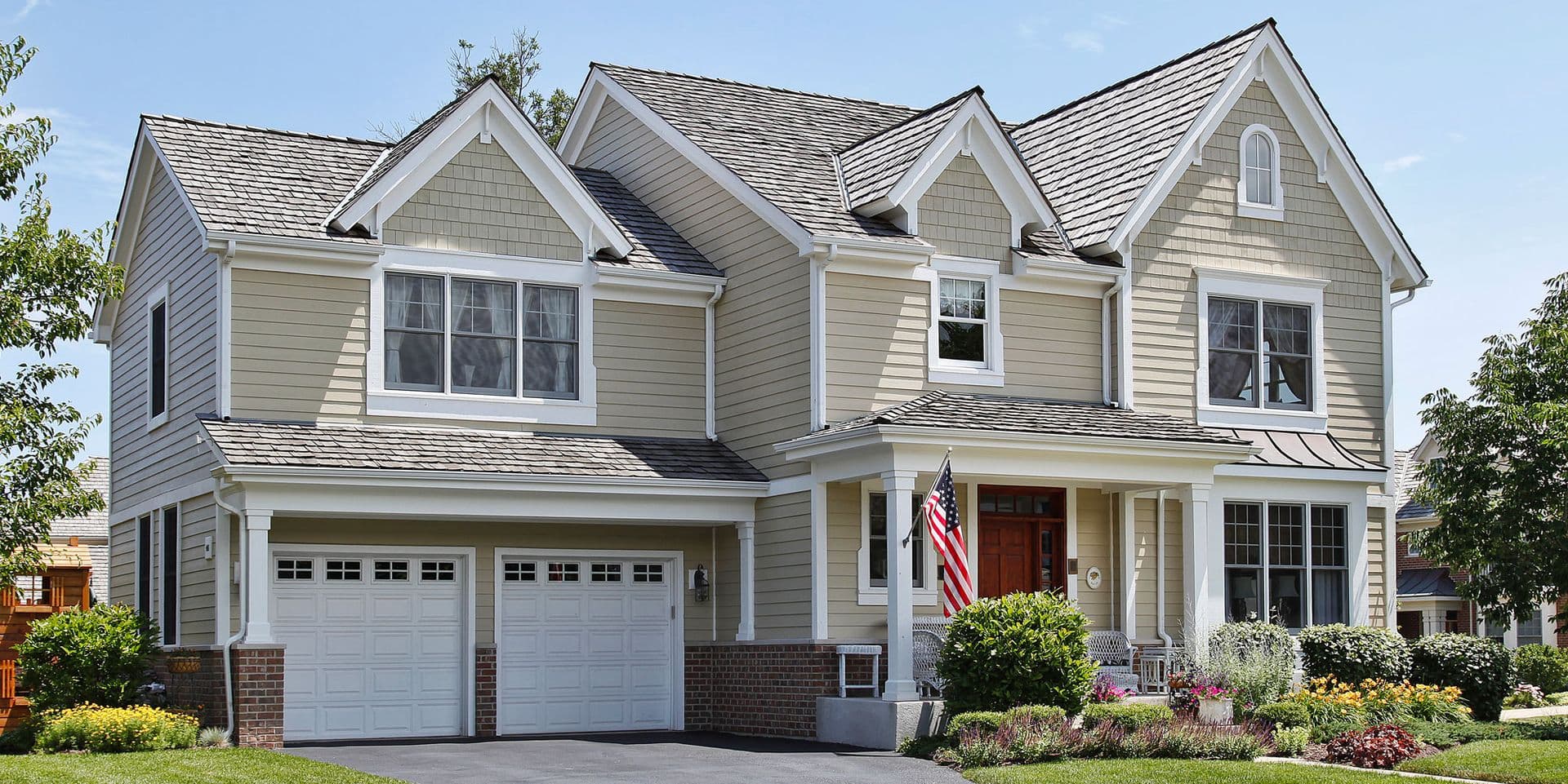 Two-story suburban home with gray siding, white trim, multiple gables, dark shingle roof, two-car garage, front porch with red door and American flag, and manicured landscaping