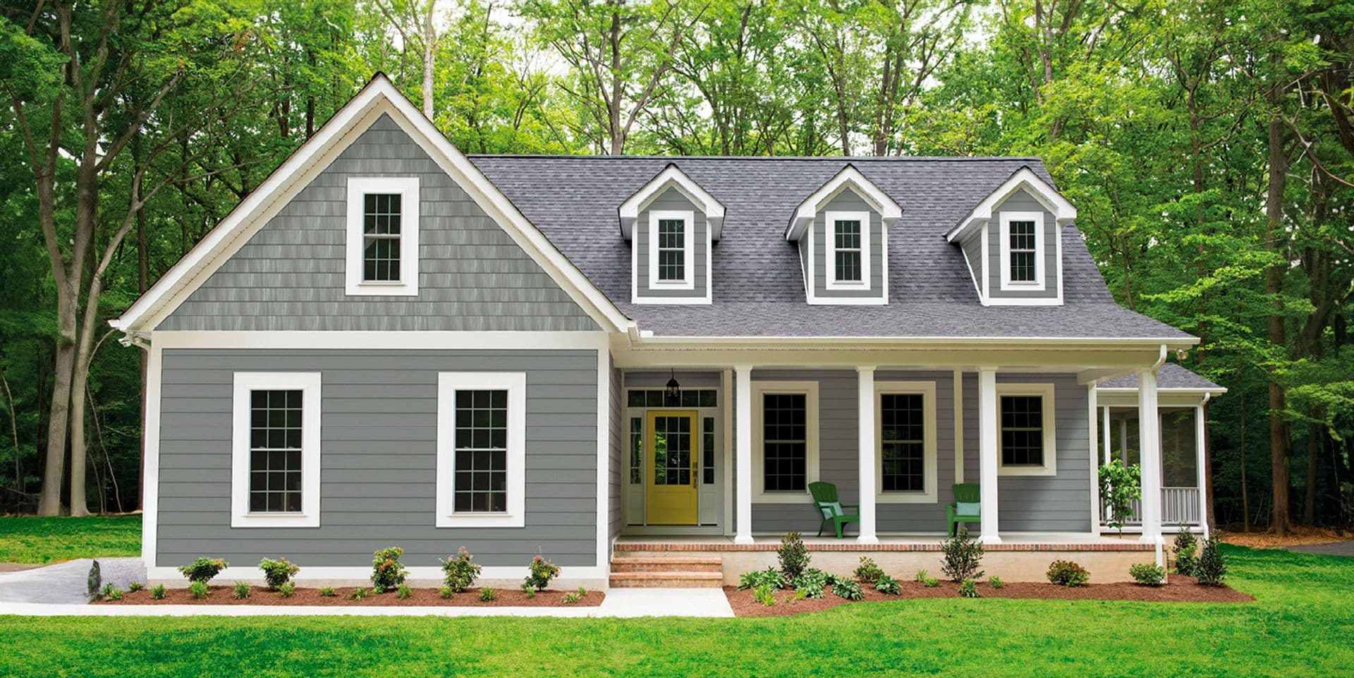 Modern farmhouse with gray siding, white trim, and dark roof set in wooded lot with front porch, dormers, and manicured landscaping