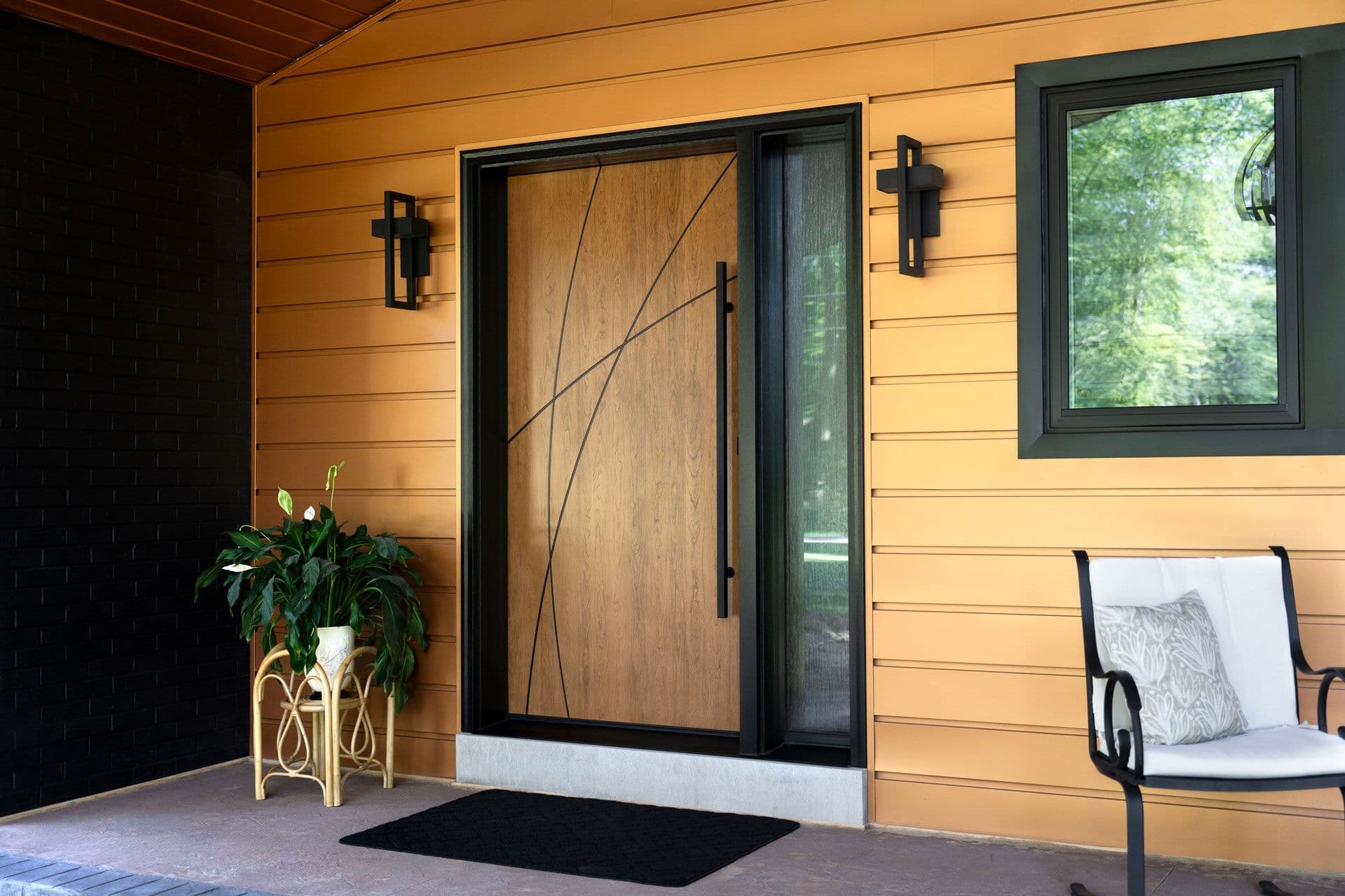 Covered porch with warm wood siding features a geometric wood entry door with black frame, decorative cross sconces, potted plant, black entry mat, and seating area with cushioned chair overlooking green landscape through window
