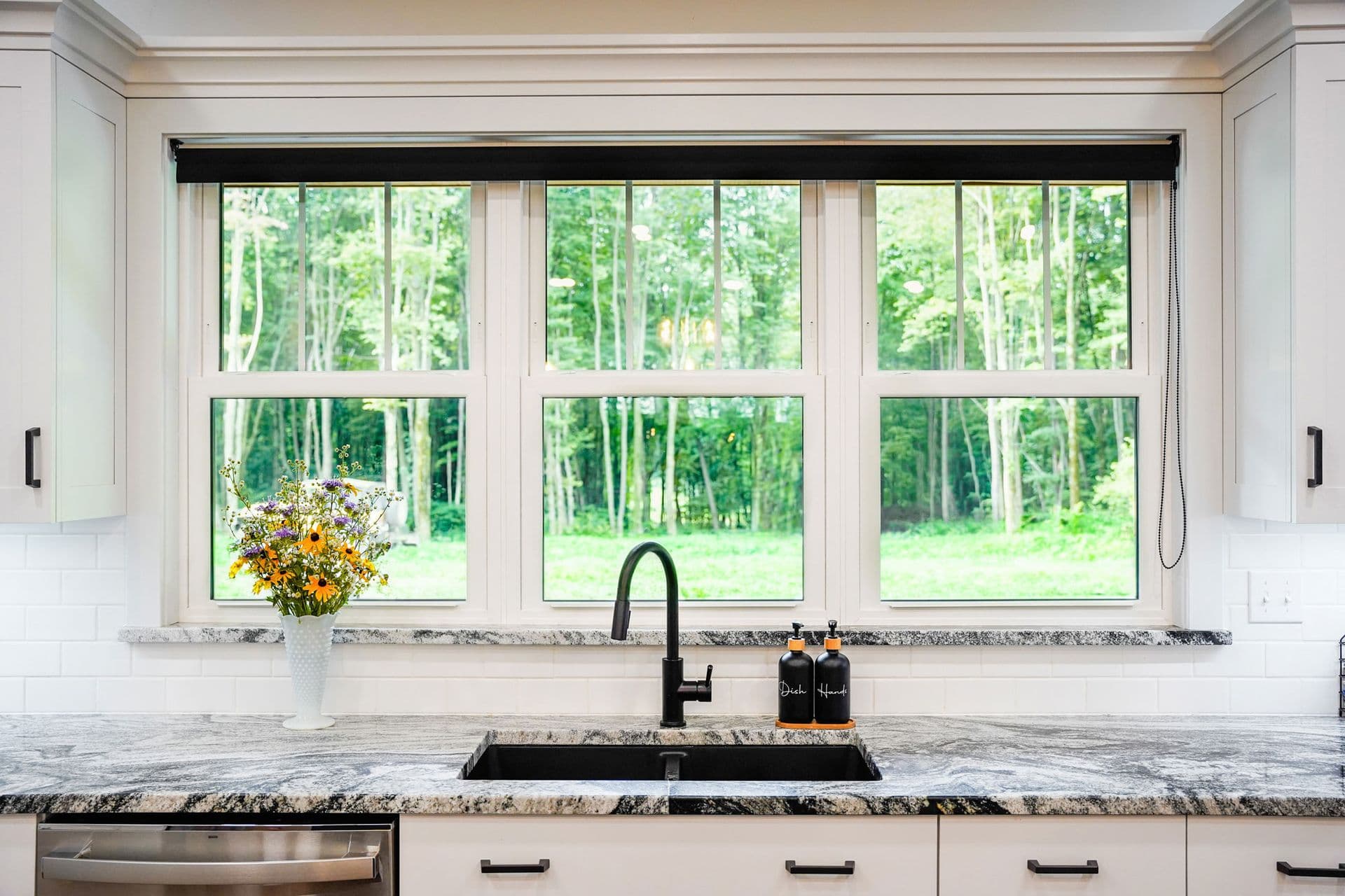 Kitchen sink with black faucet and granite countertop beneath three large windows overlooking a forest, with fresh flowers in a vase on the sill