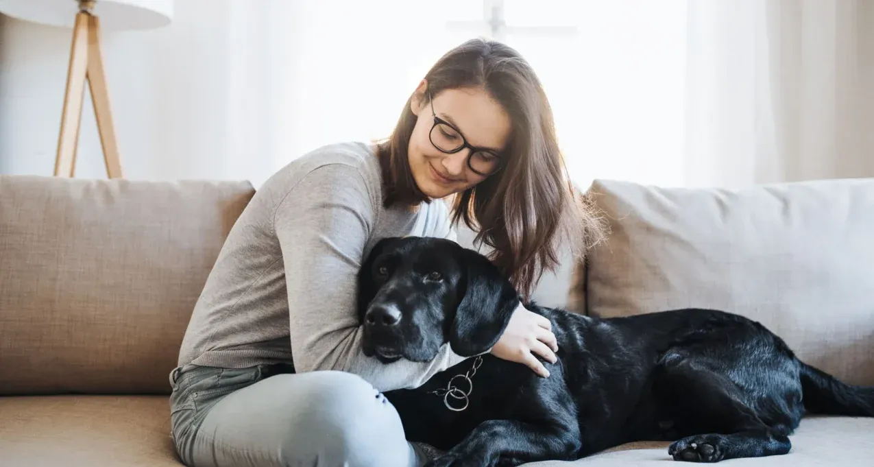 Woman wearing glasses petting a black dog on a beige sofa in a bright living room